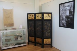 Image of a Asian inspired screen in black and gold. There is a display case on the left and a framed art work on the wall either side.